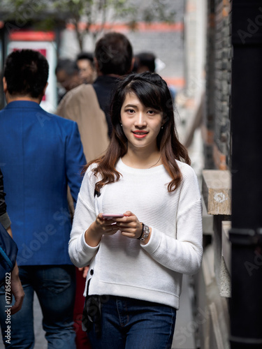 Front view of a happy Chinese girl walking and using a smart phone in crowded street.
