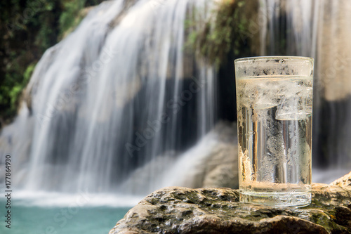 Fototapeta Naklejka Na Ścianę i Meble -  Dewy glass full of water with ice cube stands on a rock near a waterfall in the forest. A refreshing drink from nature.