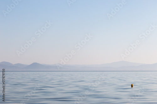 Wide and minimal view of a lake, with a small buoy on the water and distant hills, beneath a big, empty sky