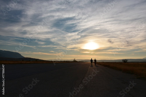 Sunset, two people walking on a wide straight empty asphalt road towards the sun at the horizon