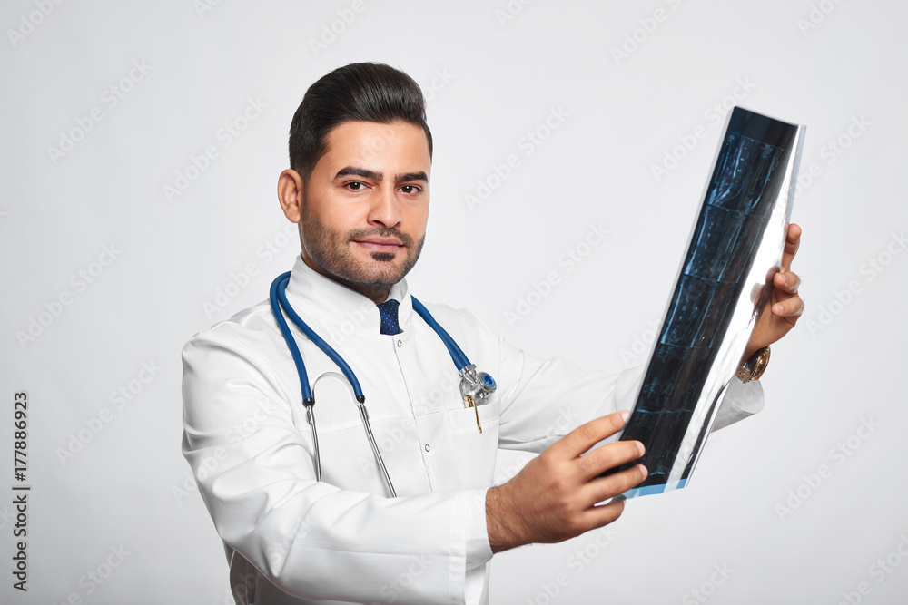 Attractive young male doctor looking to the camera seriously while examining an x-ray scan of his patient posing on grey background professionalism confidence experience healthcare help curing. Attractive young male doctor looking to the camera seriously while examining an x-ray scan of his patient posing on grey background professionalism confidence experience healthcare help curing.