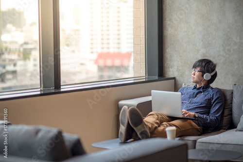 young asian man relaxing enjoy watching movie from his laptop computer with headphones on sofa, urban lifestyle in living space