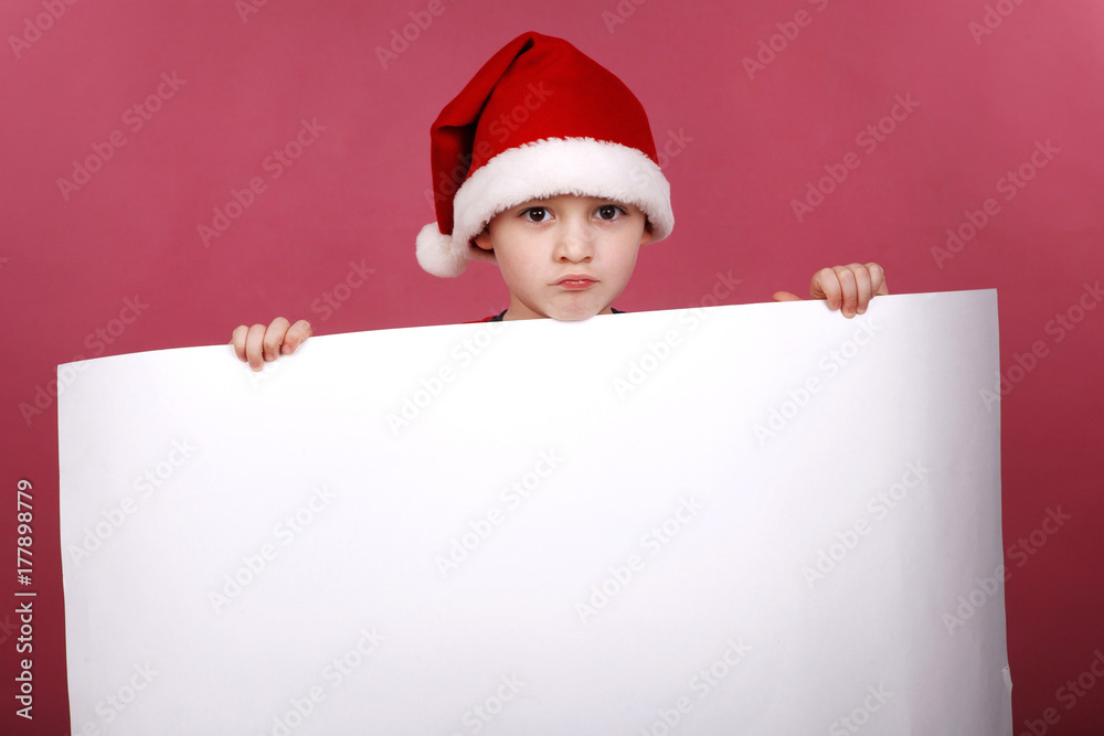 Portrait of little boy in the hat of Santa Claus on a red background , holding a white sheet of paper Portrait of little boy in the hat of Santa Claus on a red background , holding a white sheet of paper