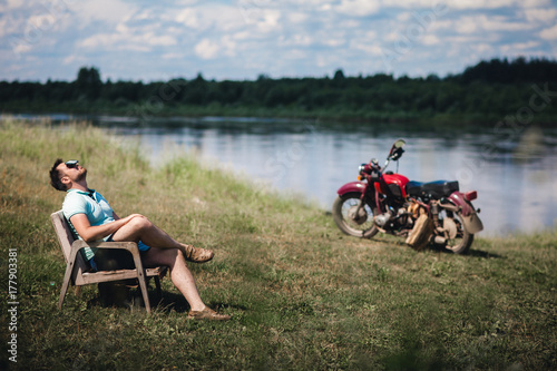 A young handsome man relaxes in an armchair against the background of a river and an old motorcycle. The concept of relaxation from modern gadgets.