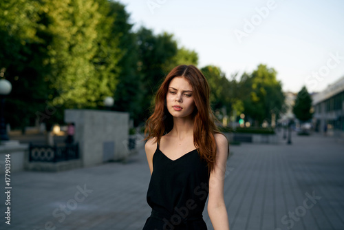A beautiful girl with red hair and a black dress strolls through the city
