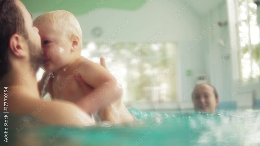 Cute baby having swimming lesson with his mother and father. Healthy family teaching their baby to swim in the swimming pool. Young father takes his son and embraces him while the kid is smiling