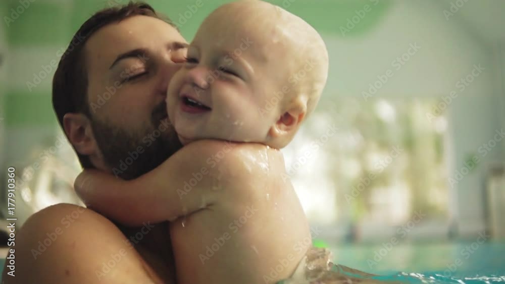 Cute little baby and his father having swimming lesson in the pool. The father is holding his son in his hands and embracing him. Little boy is happily smiling