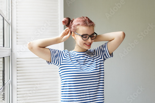 Morning routine of adorable cute pink haired teenage girl wearing sailors shirt and eyeglasses doing hair before going to school or meeting friends, smiling happily, anticipating good nice day photo