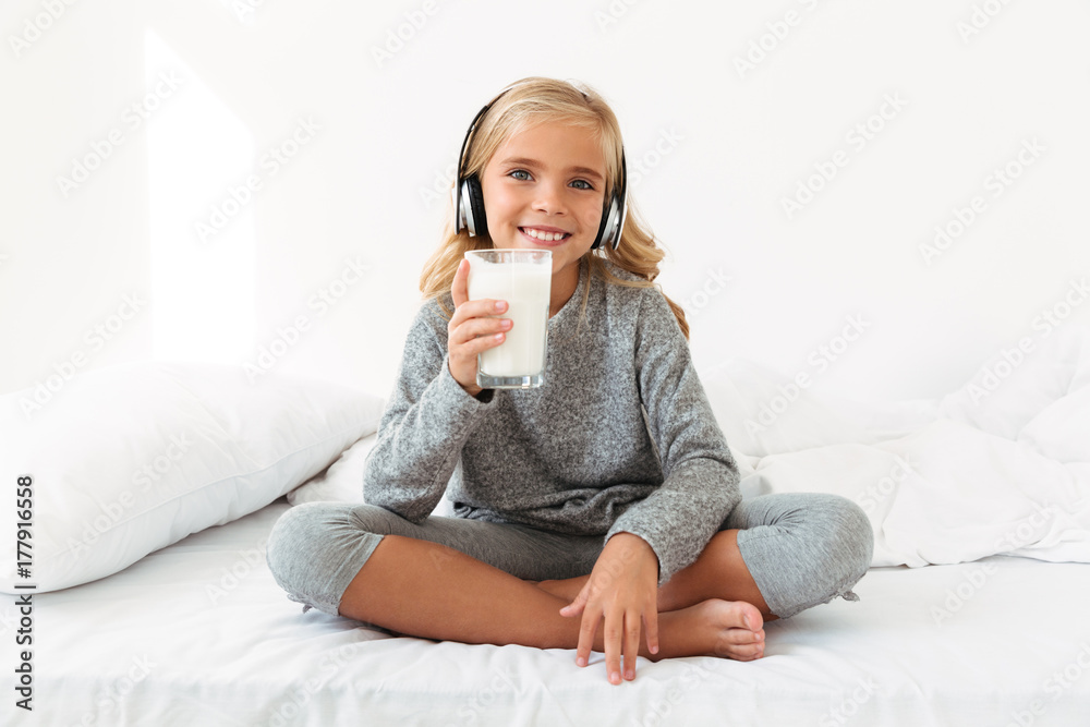 Pretty little girl in headphones holding glass of milk, sitting in bed, looking at camera Pretty little girl in headphones holding glass of milk, sitting in bed, looking at camera