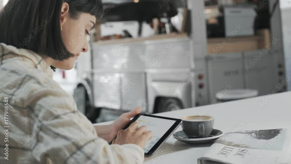 Backside view on young freelance girl is working from cafe, planning her shcedule for week and texting mails to clients on portable smartphone tablet next to morning coffee cup and newspaper Backside view on young freelance girl is working from cafe, planning her shcedule for week and texting mails to clients on portable smartphone tablet next to morning coffee cup and newspaper