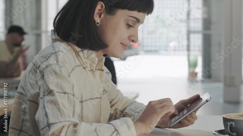 Side view on attractive smiling bunette girl using touch screen tablet to text mails or message, sitting in big spacious industrial cafe shop or coworking center photo