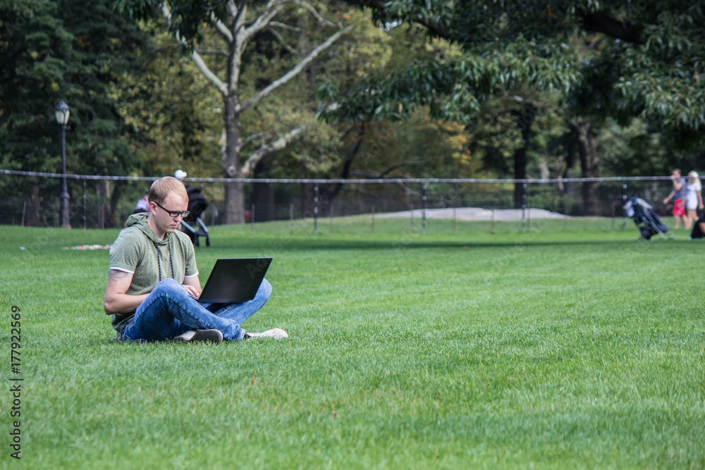 Young man in glasses work on laptop sitting on the lawn grass in park. Maybe student or hacker or businessman or startup worker