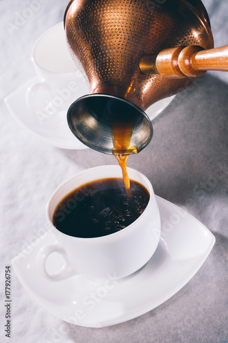 Pouring black coffee into small european white cup from old copper turkish Cezve