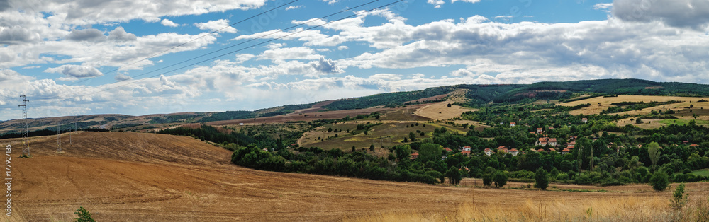A typical natural landscape of Bulgaria: agricultural fields in the hills under a cloudy sky, a village of houses with red roofs. Panoramic view.