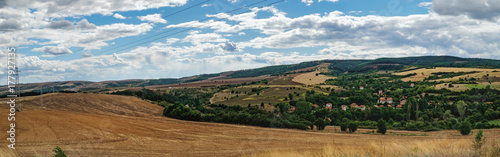 A typical natural landscape of Bulgaria: agricultural fields in the hills under a cloudy sky, a village of houses with red roofs. Panoramic view. © sonatalitravel