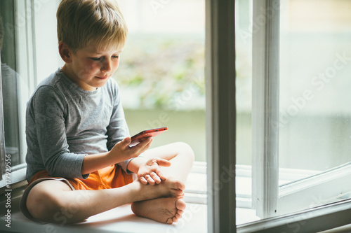 Adorable little blond kid b. oy sitting on the windowsill with rain shoes and looking on autumn raindrops, indoors. A child looks out the window. Vintage colororange rubber boots standing near window photo