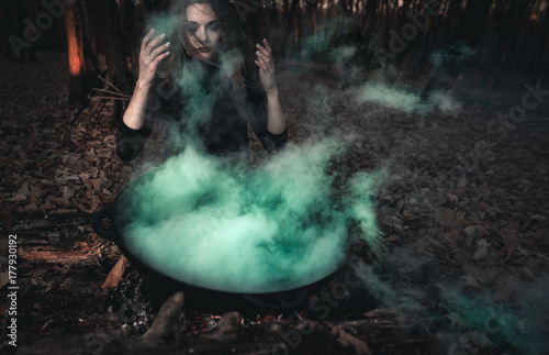 A beautiful girl witch cooking an infernal potion in a cauldron at the stake in a dark forest. The Art of Halloween