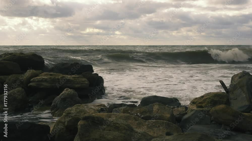 Foamy waves with a spray roll on the big rocks on the shore after a storm and the sky with clouds