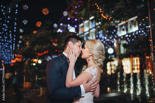 A couple of newlyweds kiss gently on the street in the evening surrounded by city lights.