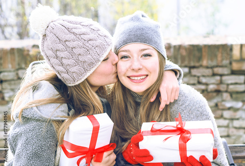 Teenagers sisters exchanging present and kissing outdoors at winter season - Best female friends hugging at park holding gift looking at camera and smiling - Concept of friendship holidays and love