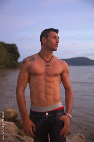 Muscle sexy man posing at the sea at colorful sunset on background