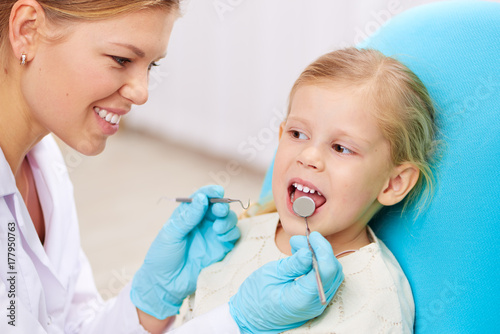 Joyful female dentist examining teeth of little girl with dental instruments. 