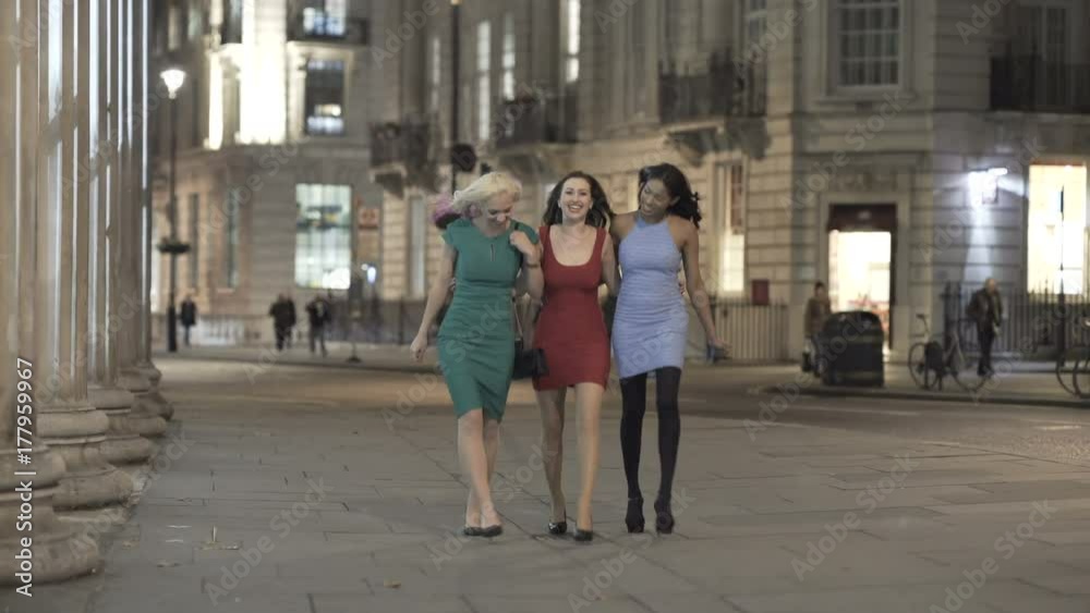  Attractive female friends in evening wear walking through the city at night.