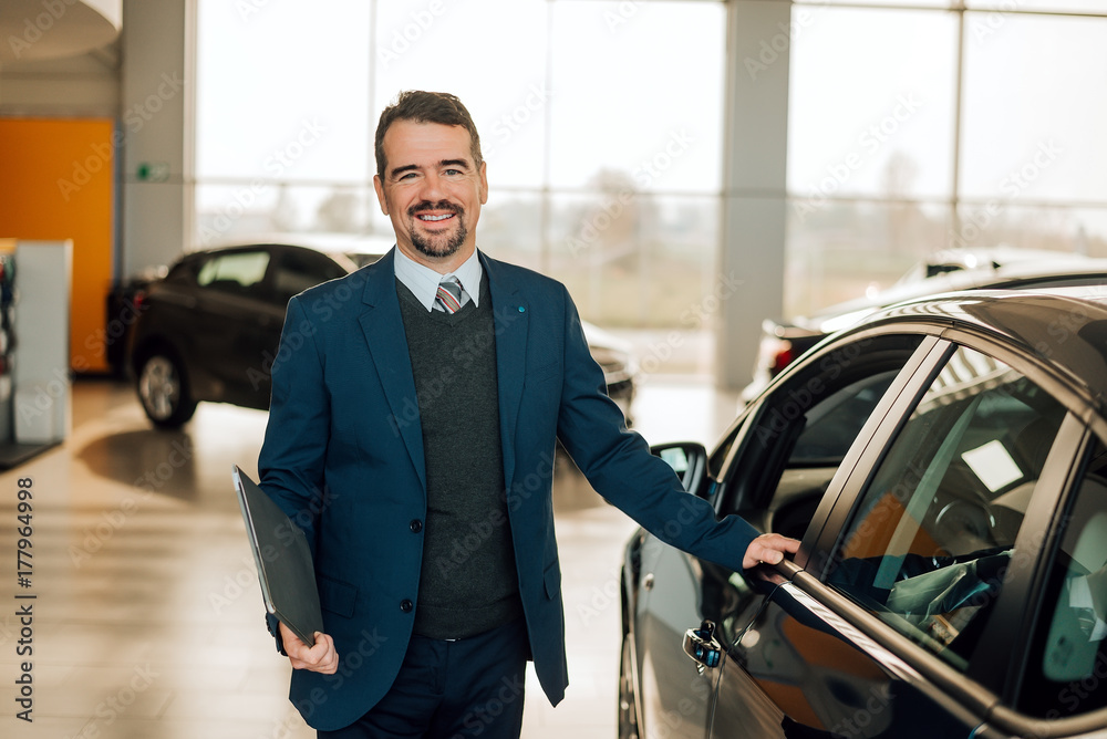 Handsome car dealership worker in suit is holding a folder and smiling while standing near the car. Handsome car dealership worker in suit is holding a folder and smiling while standing near the car.
