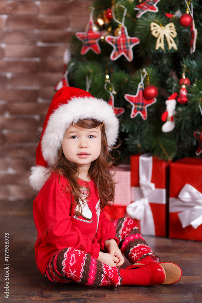 Young brunette dolly lady girl stylish dressed in red pullover sweater warm sox Santa hat shoes smiling posing sitting in studio winter christmas tree with presents with pout lips and pink cheeks. Young brunette dolly lady girl stylish dressed in red pullover sweater warm sox Santa hat shoes smiling posing sitting in studio winter christmas tree with presents with pout lips and pink cheeks.