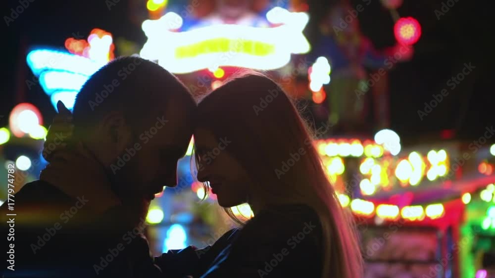 beautiful woman and bearded man staying at night head to head hugging together dancing in theme park with multicoloured lights of rides background slow motion closeup beautiful woman and bearded man staying at night head to head hugging together dancing in theme park with multicoloured lights of rides background slow motion closeup