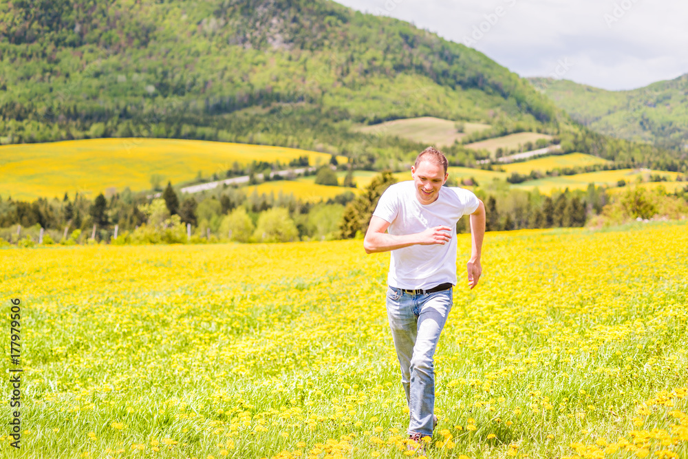 Young man running, jumping in air and smiling on countryside yellow dandelion flower fields in summer grass in Ile D'Orleans, Quebec, Canada happy
