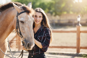 Portrait of posh and beautiful long haired young woman taking care about a horse on a ranch. Sunny summer day outside.