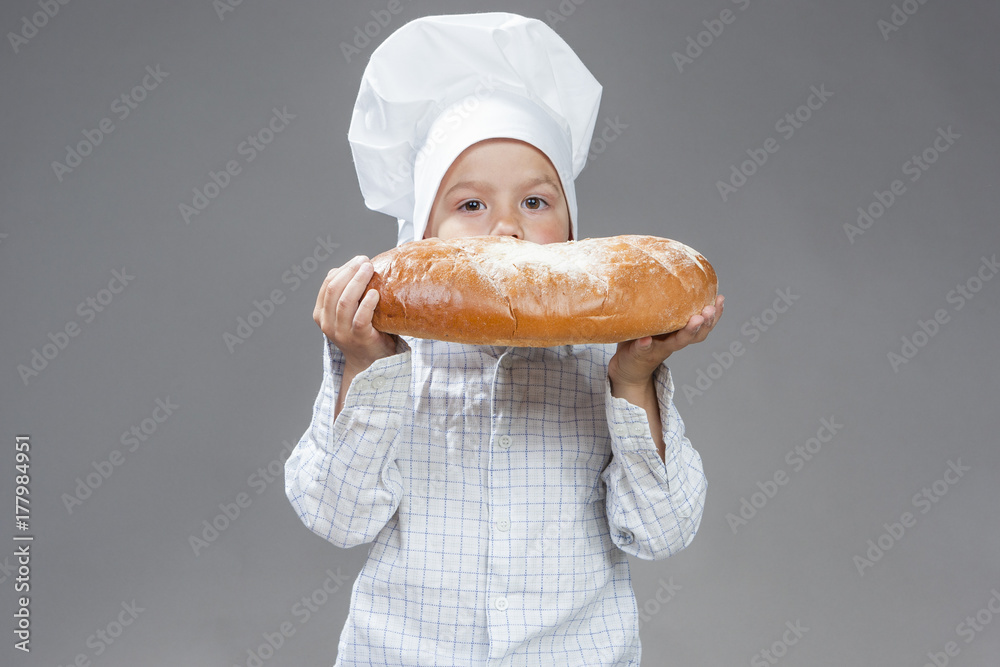 Food Concepts. Portrait of Happy Caucasian Little Boy Eating Big Fresh Bun with Sugar. Posing In Cooking Hat Agaunst Gray Background. Food Concepts. Portrait of Happy Caucasian Little Boy Eating Big Fresh Bun with Sugar. Posing In Cooking Hat Agaunst Gray Background.