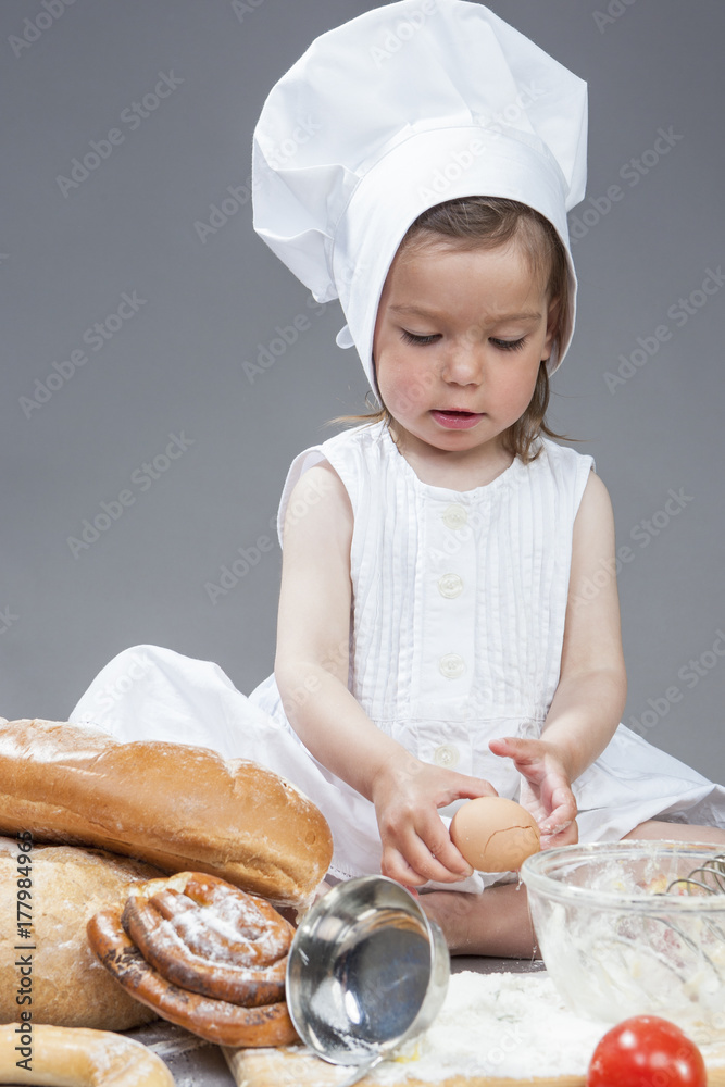 Caucasian Girl In Specific Cook Uniform Breaking Down Fresh Egg and Making Food in Kitchen Glassware with Whisk In Studio Environment. Against Gray Background. Caucasian Girl In Specific Cook Uniform Breaking Down Fresh Egg and Making Food in Kitchen Glassware with Whisk In Studio Environment. Against Gray Background.