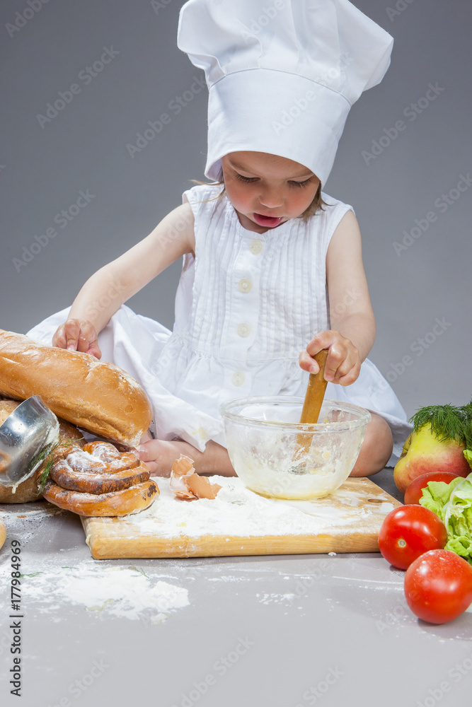 Cooking Ideas. Caucasian Little Girl In Cook Uniform Working With Whisk and Kitchen Glassware In Studio Environment. With Vegetables and Fruits on Background. Cooking Ideas. Caucasian Little Girl In Cook Uniform Working With Whisk and Kitchen Glassware In Studio Environment. With Vegetables and Fruits on Background.