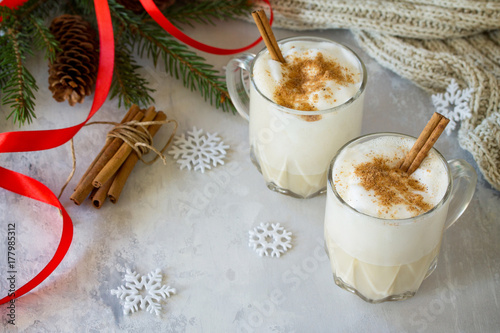 Homemade Eggnog Christmas milkshake with cinnamon, served in two cups on a gray stone or slate background, decorative ornaments, Christmas tree with copy space.