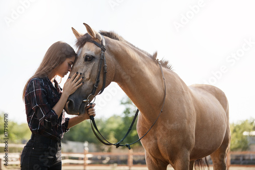 Portrait of young female assistant farm manager calming down a horse and talking to a new stallion. © Liubov Levytska