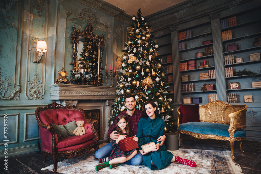 Friendly family sits near decorated Christams tree, embrace each other, hold packed presents, enjoy majestic atmospere. Father, mother and daughter near New Year tree in living room. Festive concept