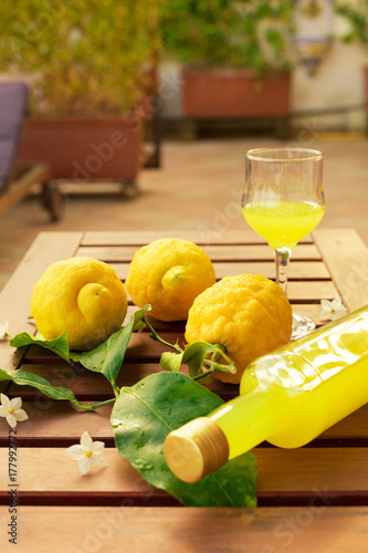 Quadro su tela Lemonade or limoncello in a glass bottle, glasses, lemons with leaves on a wooden table on the terrace