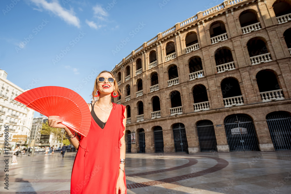 Portrait of a woman in red dress with spanish hand fan stnading in front of the bullring amphitheatre in Valencia city, Spain