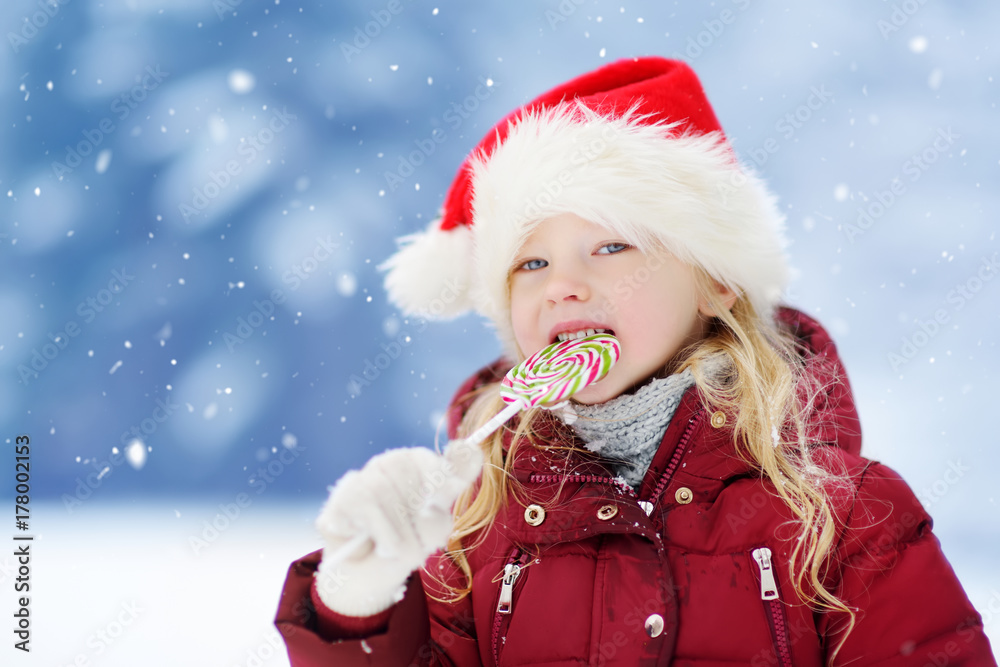 Adorable little girl wearing Santa hat having huge striped Christmas lollipop on beautiful winter day Adorable little girl wearing Santa hat having huge striped Christmas lollipop on beautiful winter day