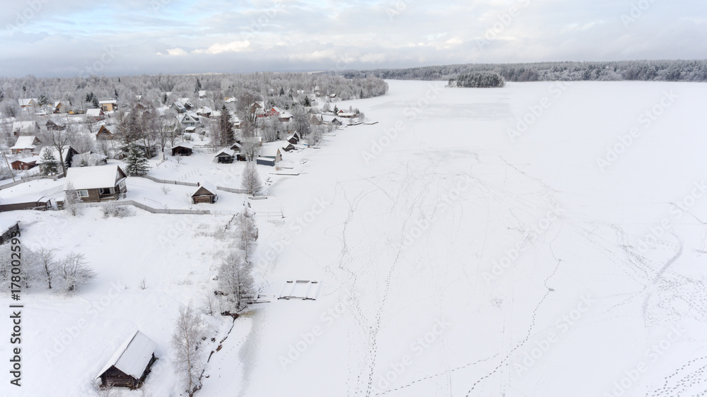 Footprints are on ice of frozen lake near winter shore. Russian remote village is in evergreen forests. Aerial view. Russia Footprints are on ice of frozen lake near winter shore. Russian remote village is in evergreen forests. Aerial view. Russia