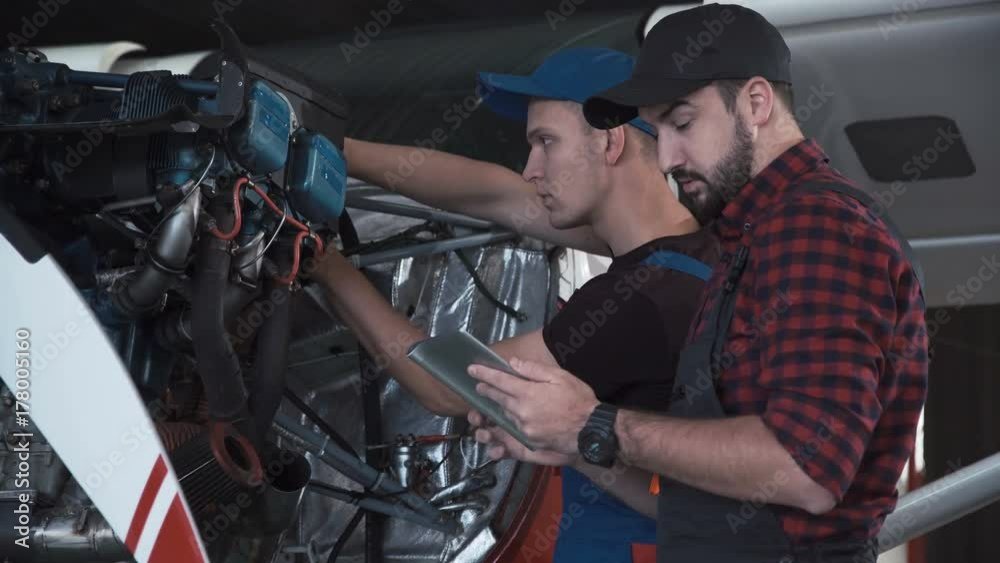 Two flight mechanics doing a pre flight check or maintenance on a small single engine aircraft using digital tablet in a hangar in a close up view of them working on the engine.
