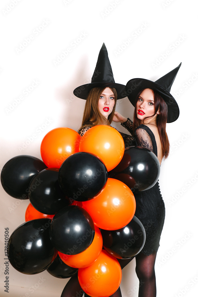 Two happy sexy women in black witch halloween costumes with orange and black balloon on party over white background