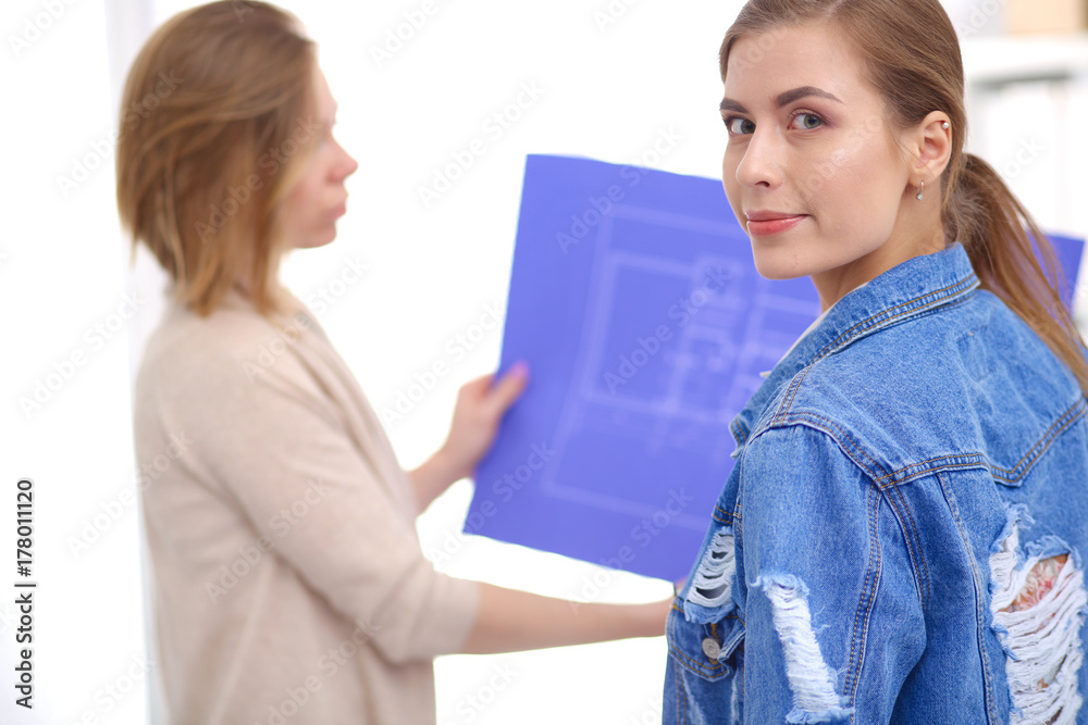 Two young woman standing near desk with instruments, plan and laptop.