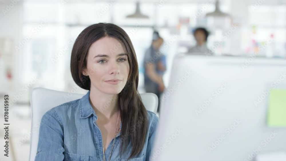 Close up young woman working on computer in creative office with colleagues talking in background. Shot on RED Epic. Close up young woman working on computer in creative office with colleagues talking in background. Shot on RED Epic.