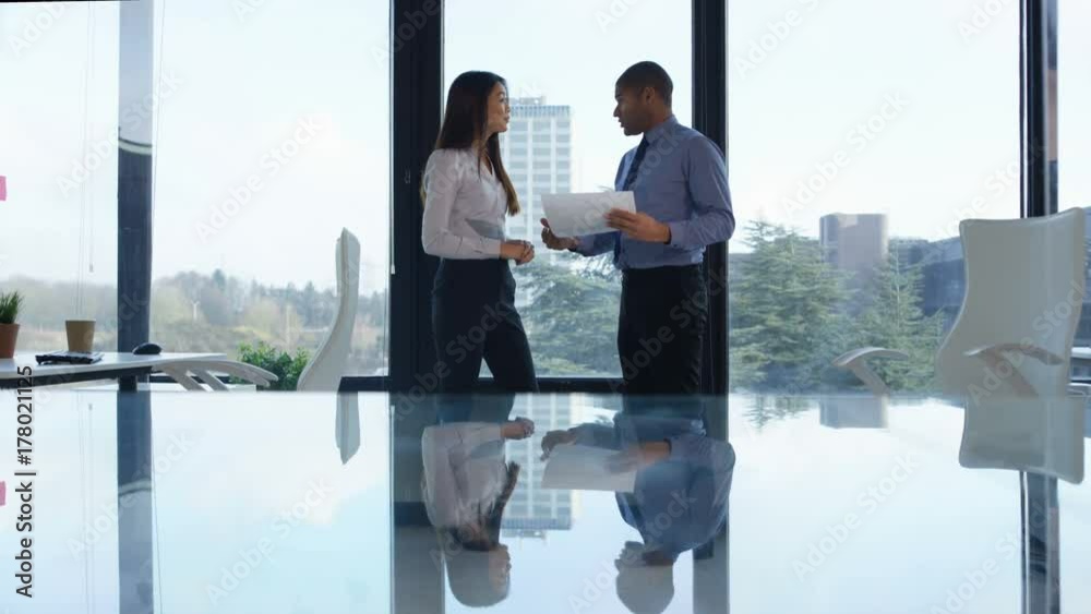 Business man & woman discussing paperwork in front of large office window Business man & woman discussing paperwork in front of large office window