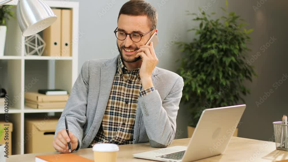 Close up shot of a cheerful man in glasses sitting at the office desk, talking on the phone and holding a pen in his hands. Indoor shot. Close up shot of a cheerful man in glasses sitting at the office desk, talking on the phone and holding a pen in his hands. Indoor shot.
