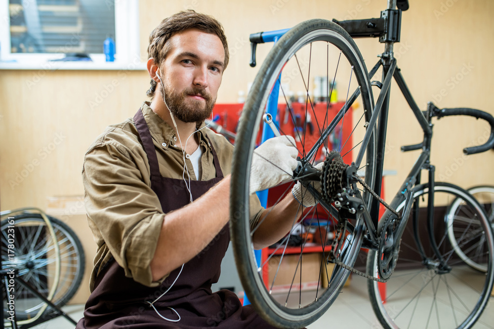 Portrait of bearded young repairman sitting on haunches and looking at camera while repairing bicycle at spacious workshop Portrait of bearded young repairman sitting on haunches and looking at camera while repairing bicycle at spacious workshop