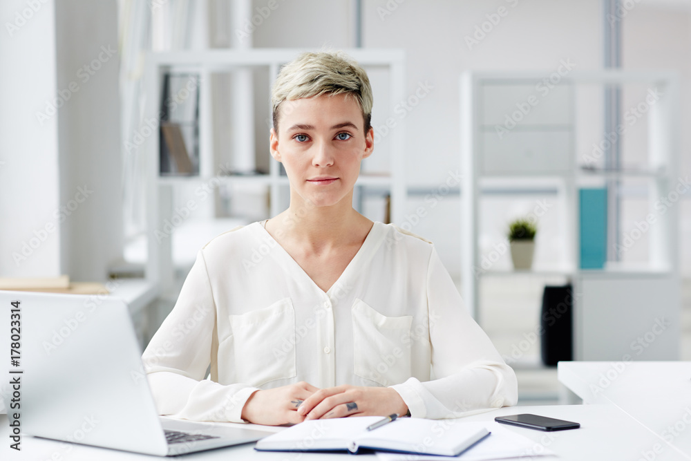 Serious young businesswoman sitting by workplace in office Serious young businesswoman sitting by workplace in office
